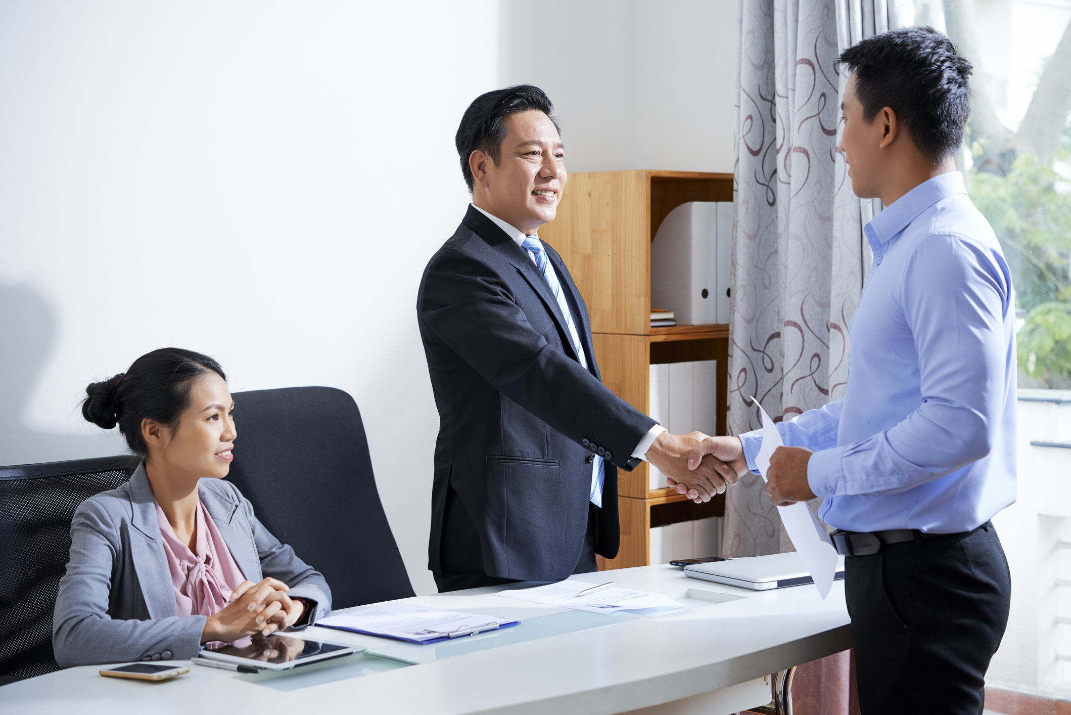 Two businessmen holding hands while a businesswoman sitting in an office