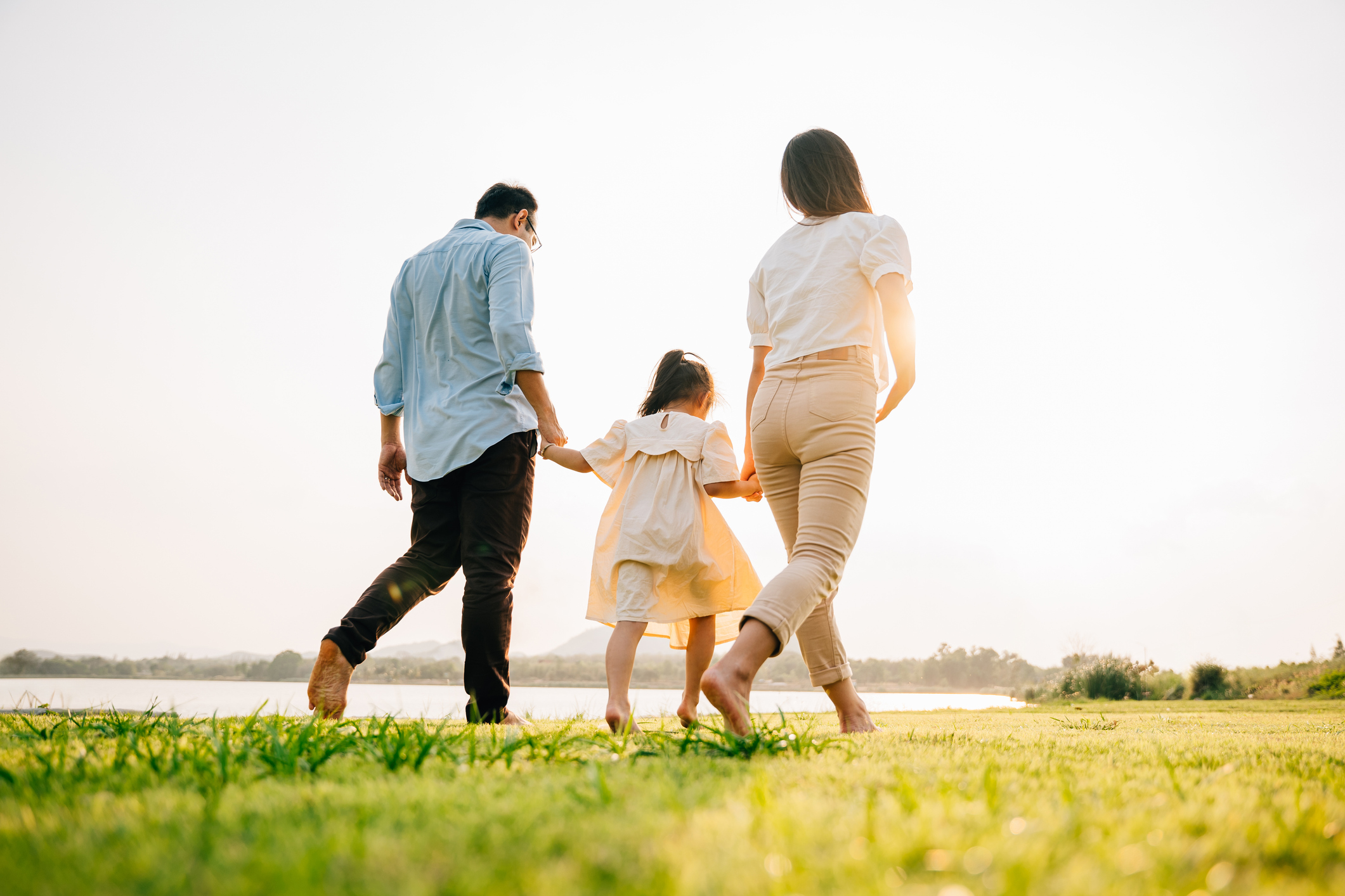 Father and mother holding daughter’s hand on a grass field by a lake