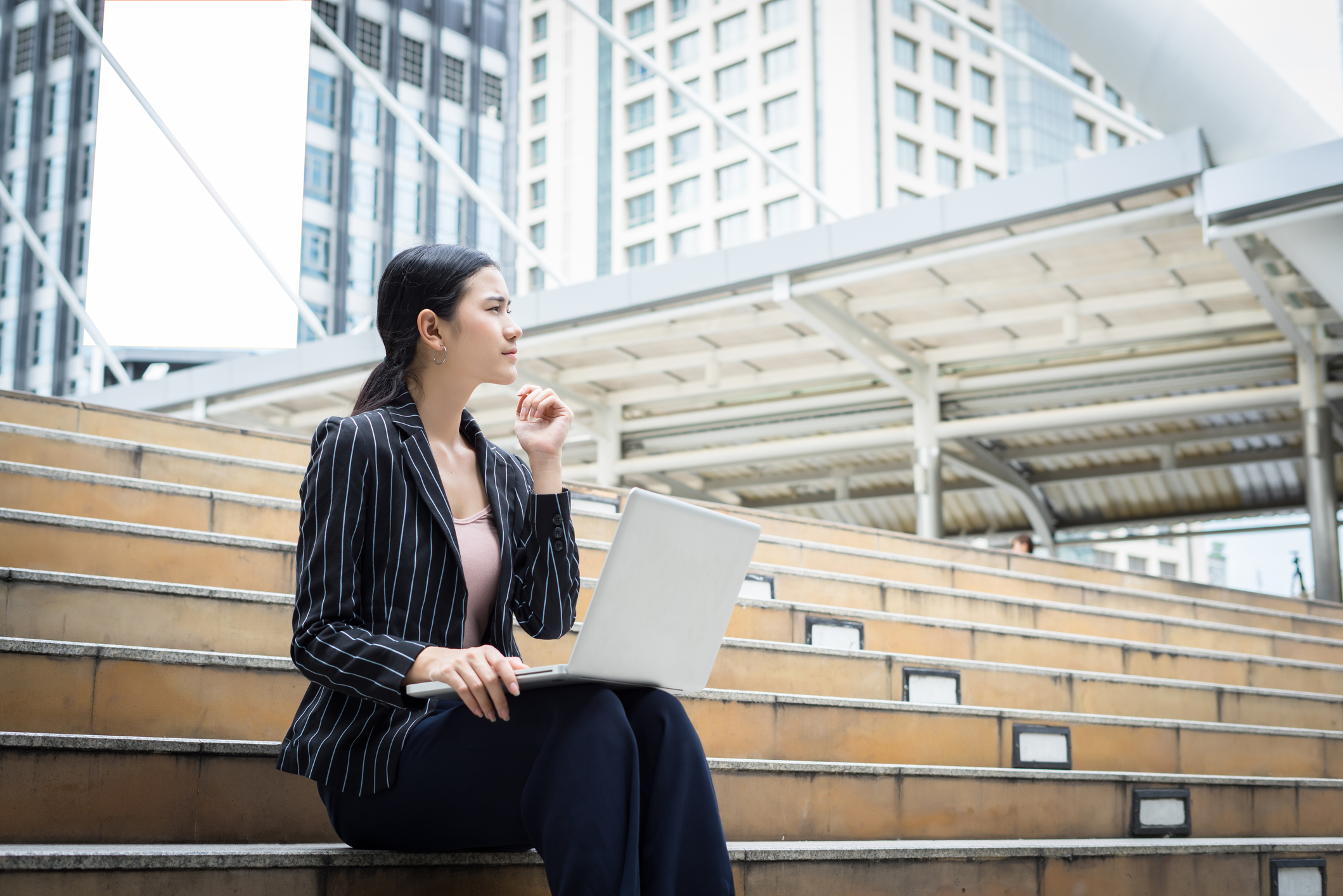 Woman sitting on a stair in an outdoor space and thinking with a laptop on her legs