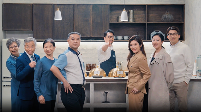Group of people standing in a kitchen and smiling while the man standing in the middle pointing his finger in front