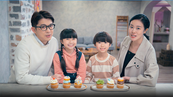 Man, woman, young girl and young boy sitting by the table with two dishes of cupcake on the table in front of them