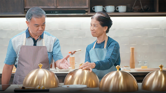 Old man and woman standing in the kitchen and ready to open the dish cover on the table