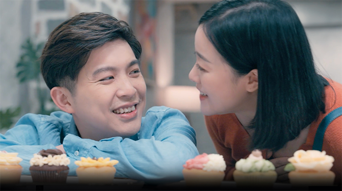 Close-up of a man and woman smiling and looking at each other with a table of cupcakes in front of them